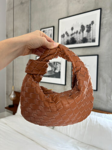 woven handbag, vegan leather knot bag, designer-inspired handbag, brown crossbody bag, luxury handbag for less, mini shoulder bag, boho chic purse, trendy vegan handbag, cruelty-free fashion, fashionable handbags 2025, Caramel brown vegan leather woven knot handbag with a chunky braided handle, held in hand against a modern bedroom background.