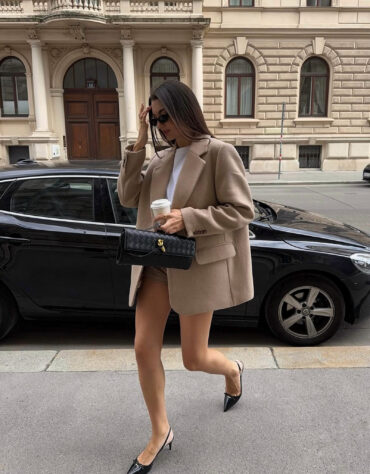 Stylová žena v béžovém oversize saku, s černými slingback podpatky a slunečními brýlemi, drží v ruce černou prošívanou kabelku z veganské kůže se zlatým zapínáním, elegantní street style outfit ideální do města.